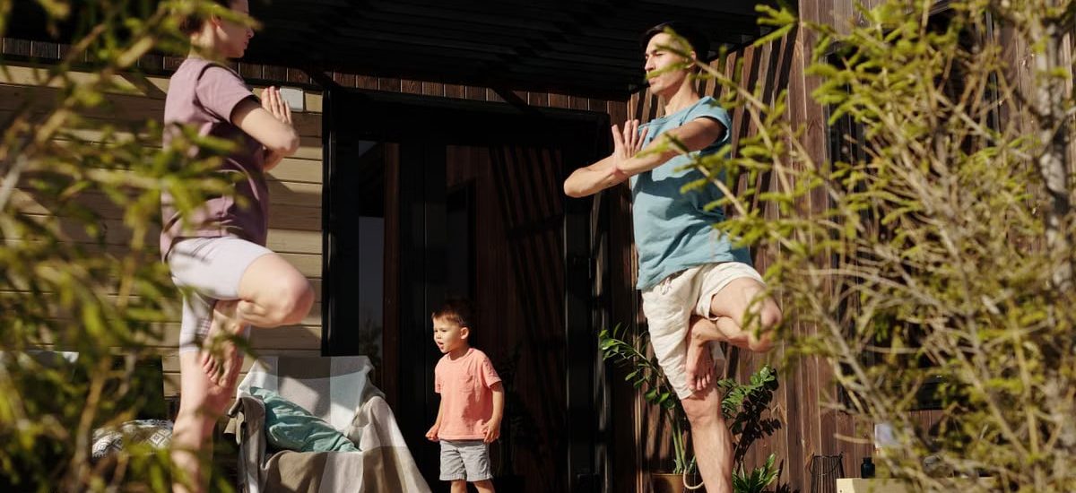 A family practicing yoga in tree pose on a sunlit porch, promoting health and mindfulness.